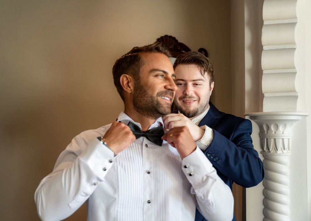 A groom adjusts his bowtie while getting ready in formal attire near a decorative white pillar.