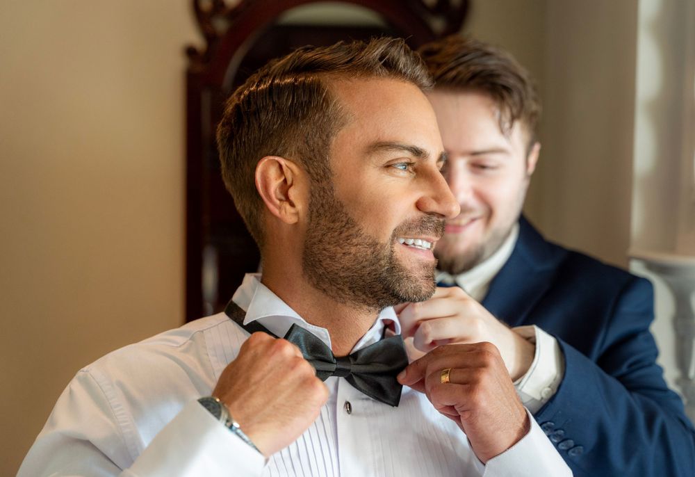Two people in formalwear share a tender moment while adjusting a bowtie by a window.
