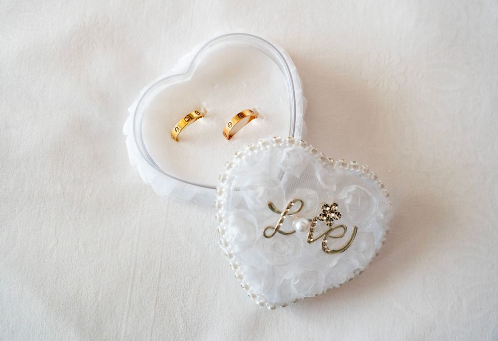 Gold wedding rings displayed in a delicate white lace heart-shaped cushion with 'Love' embroidery decoration.