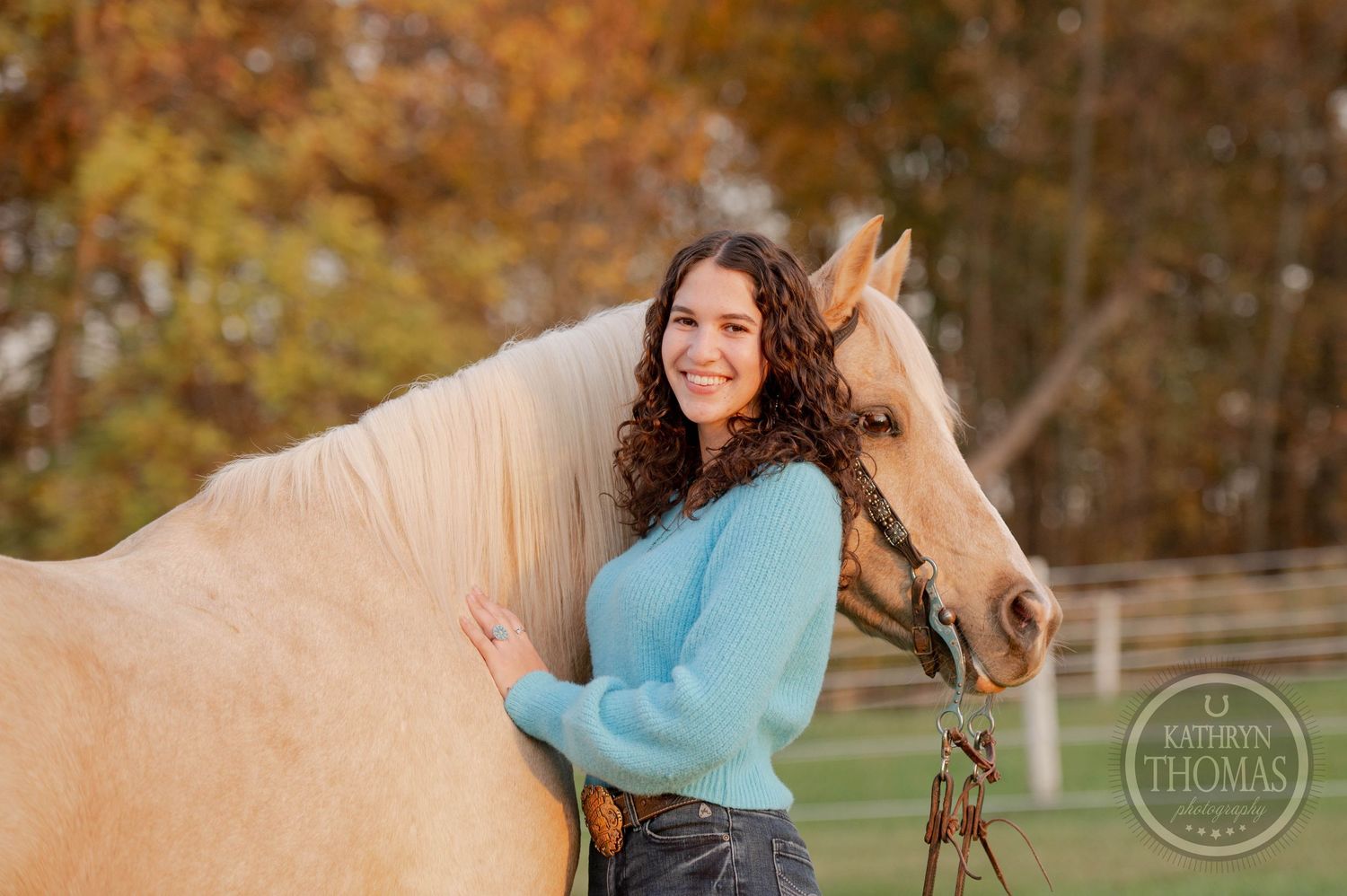 A person wearing a blue sweater poses with a palomino horse against autumn foliage.