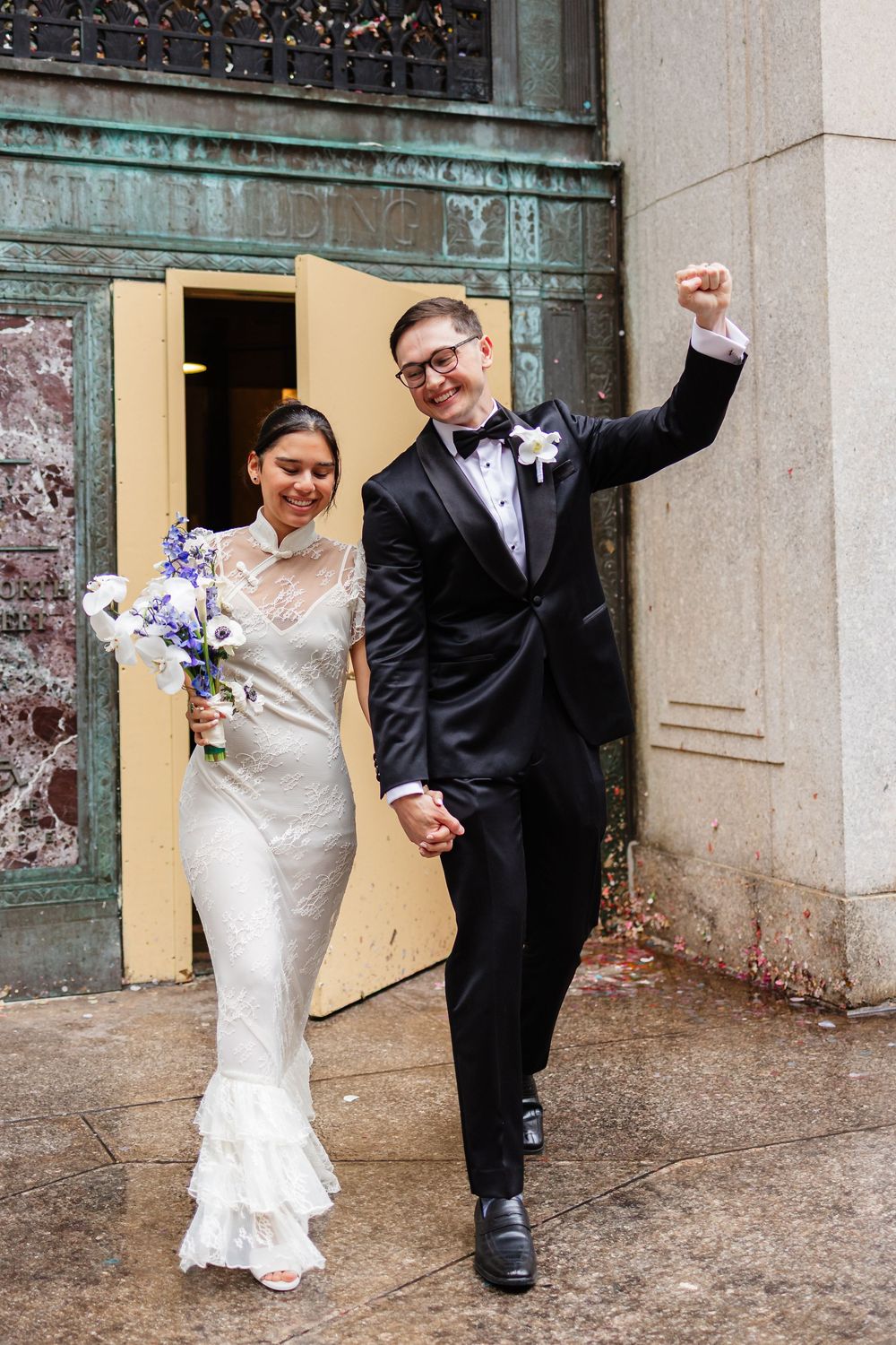 A joyful bride and groom celebrate together in front of a rustic building wall with purple floral bouquet.