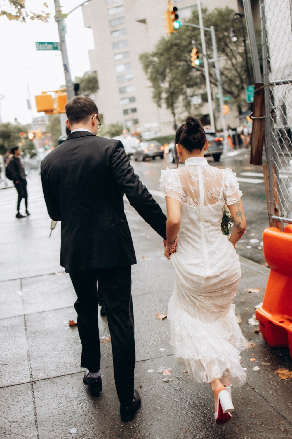 A couple in formal attire walks together on a rainy city sidewalk with traffic lights in the background.