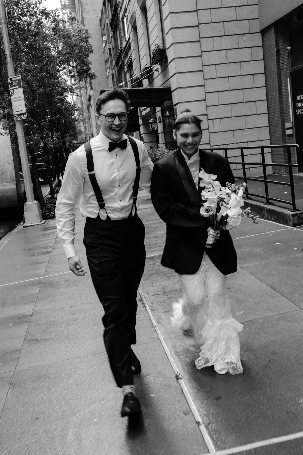 A couple in wedding attire walks together on a city sidewalk in black and white photography.