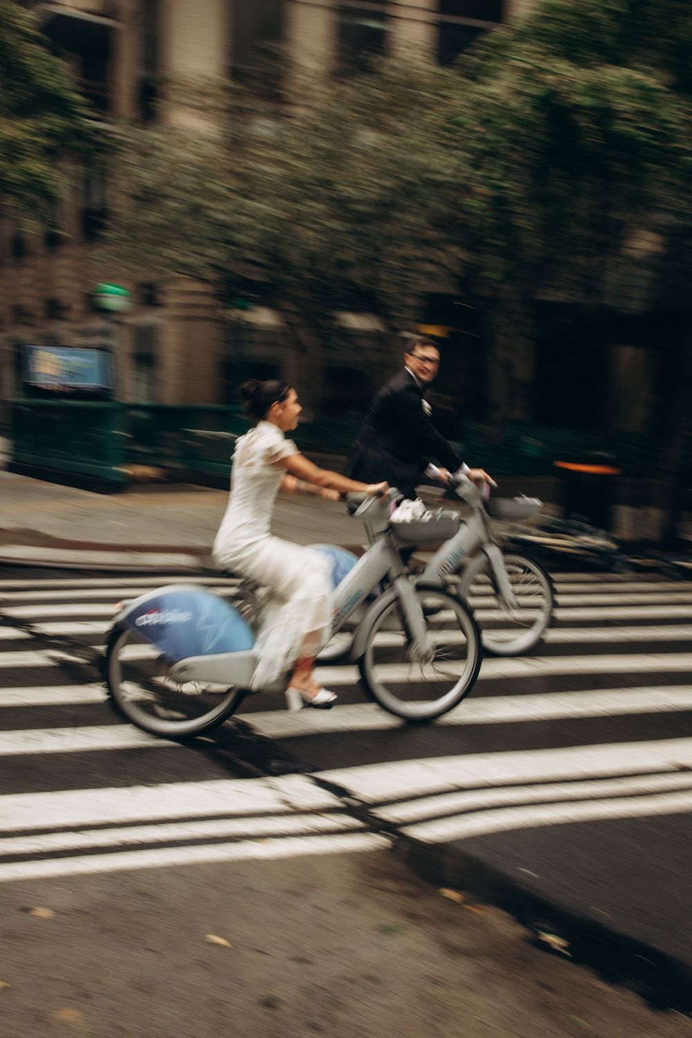 Motion blur sequence shows a cyclist in white clothing leaning forward while riding across a crosswalk in an urban setting.