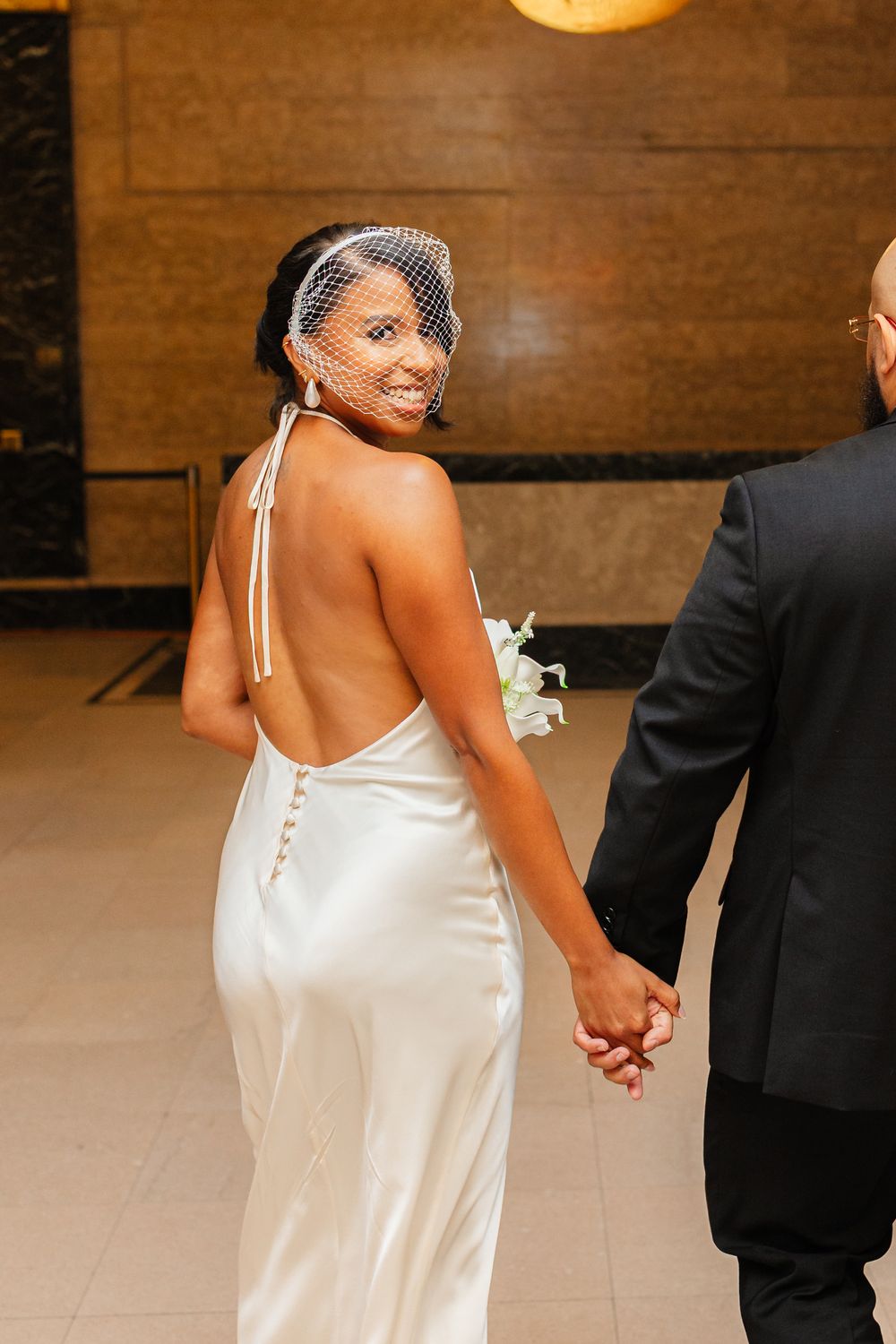 A bride in a white satin gown holds hands with her partner during a wedding ceremony in a rustic venue.