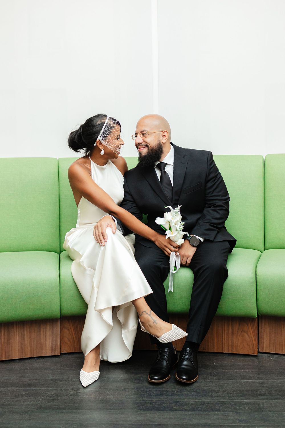 A romantic wedding couple shares a tender moment while sitting on a modern green couch in a minimalist setting.