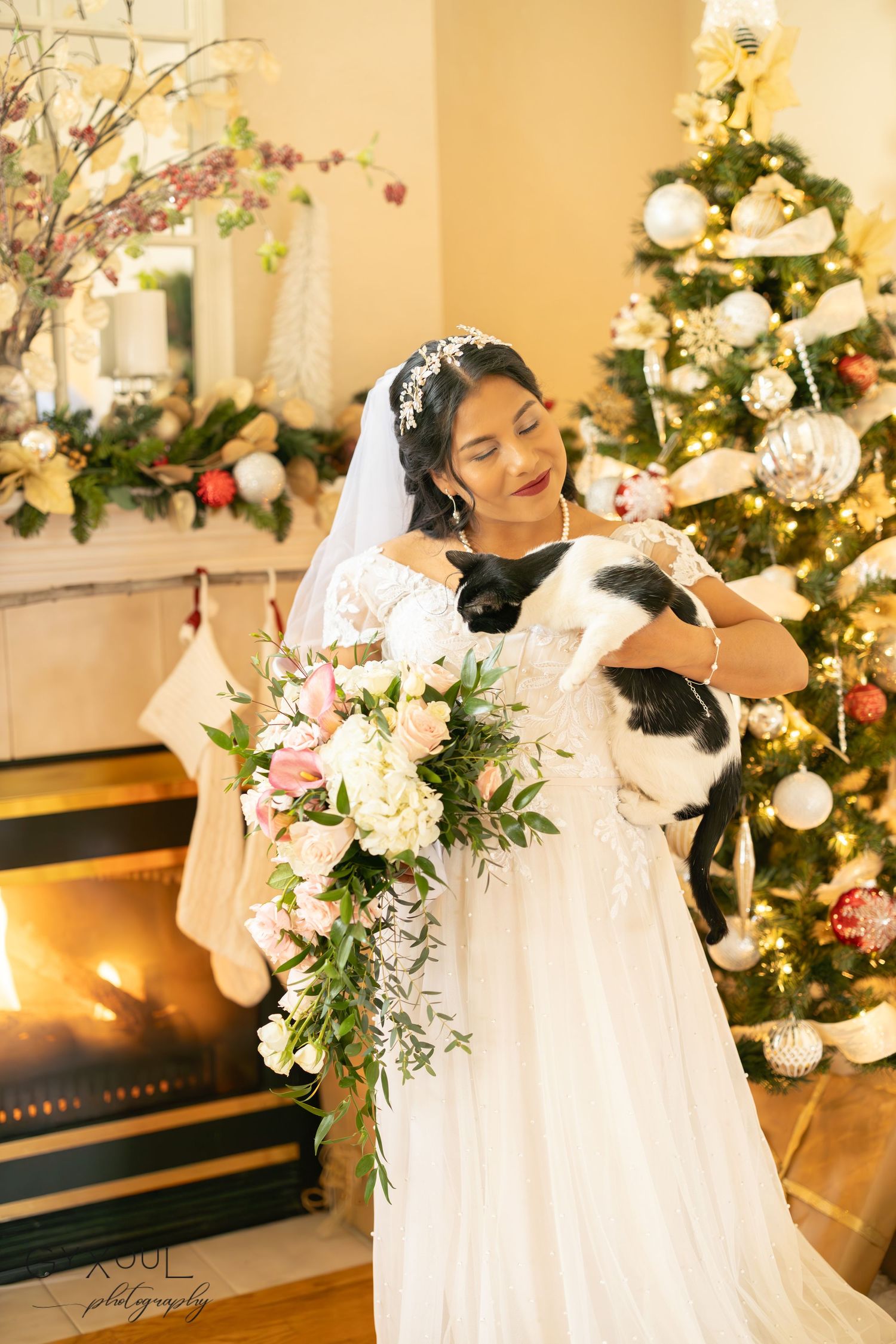 A bride in a white gown poses with her bouquet in front of a decorated Christmas tree and fireplace.