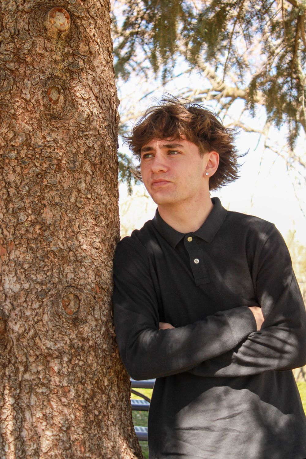 Person leaning against large tree trunk wearing dark polo shirt in natural lighting.