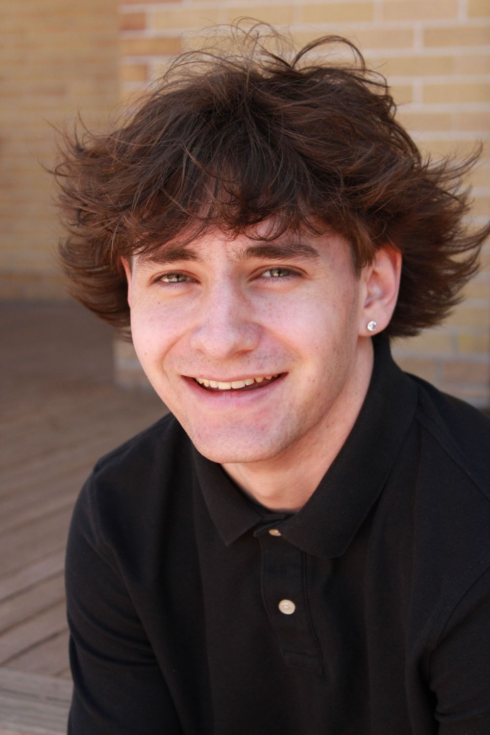 Close-up portrait showing person in black polo with tousled hair against brick wall.