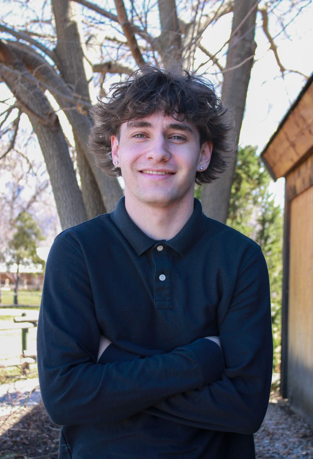 Person wearing a dark polo shirt smiling with crossed arms outside near trees.
