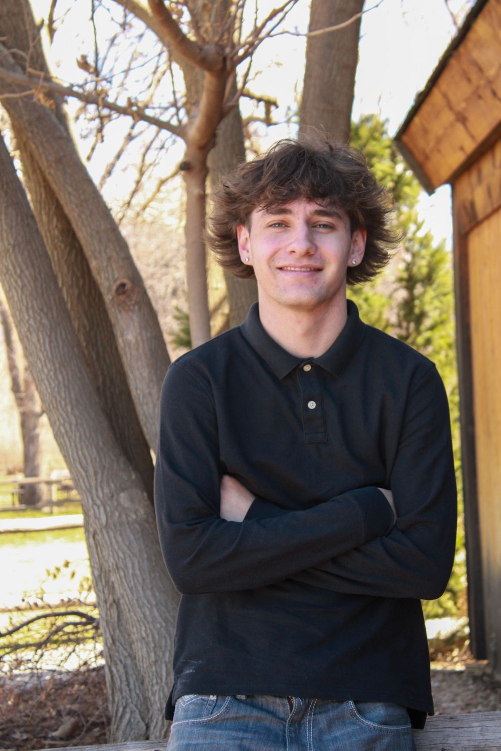 Casual outdoor portrait with someone in black polo against trees and wooden structure.