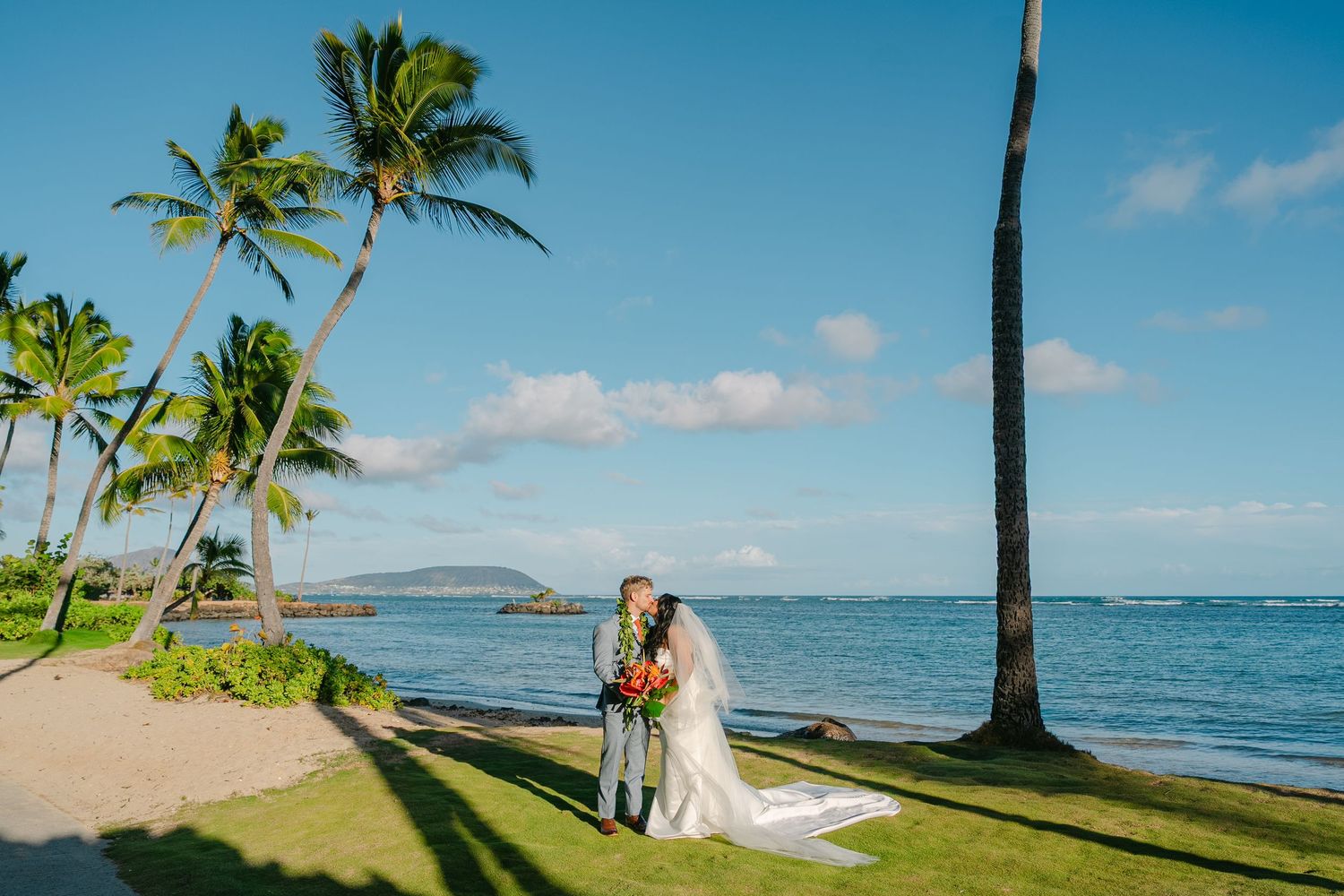 A couple walks barefoot on a tropical beach holding hands while bride holds a colorful bouquet of flowers.