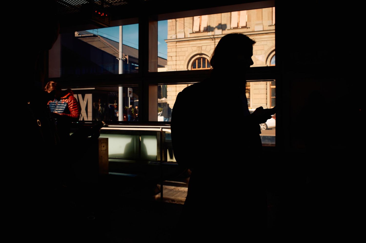 A silhouetted figure stands in front of a window with urban architecture visible outside in dramatic lighting.
