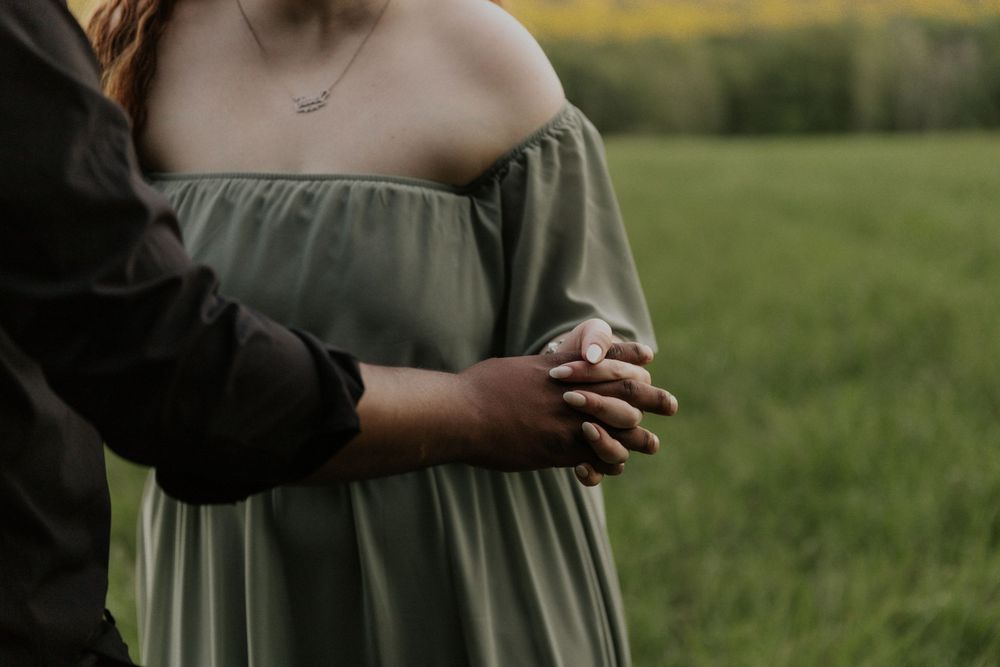 Two people stand close together in an open meadow with rolling hills in the background.