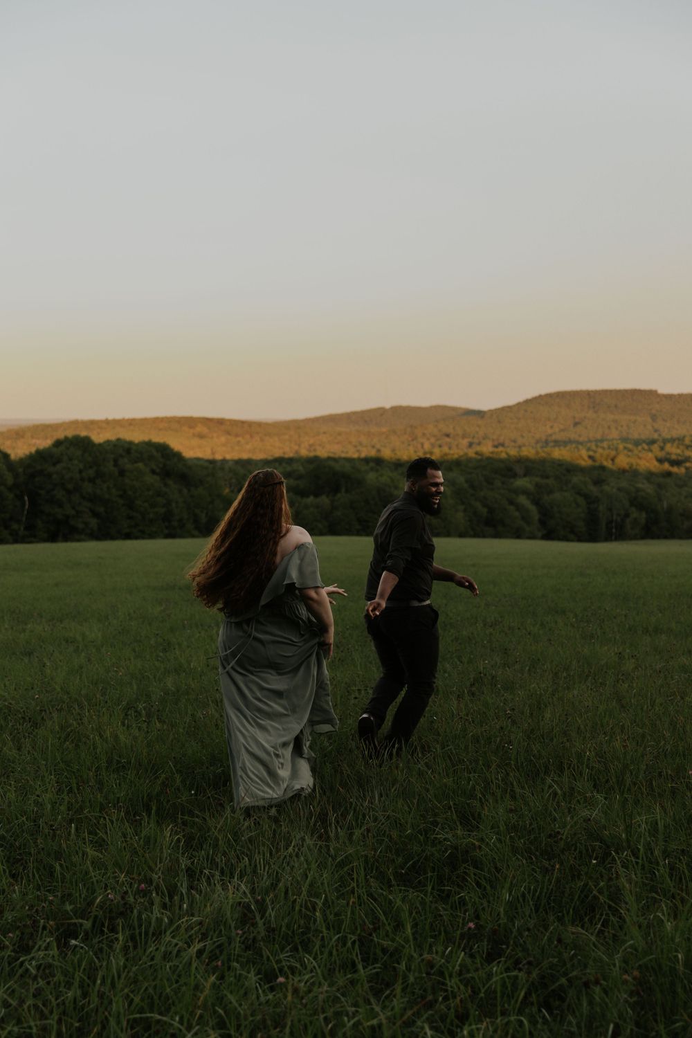 A couple dances freely in a grassy field with mountains and forest in the background.