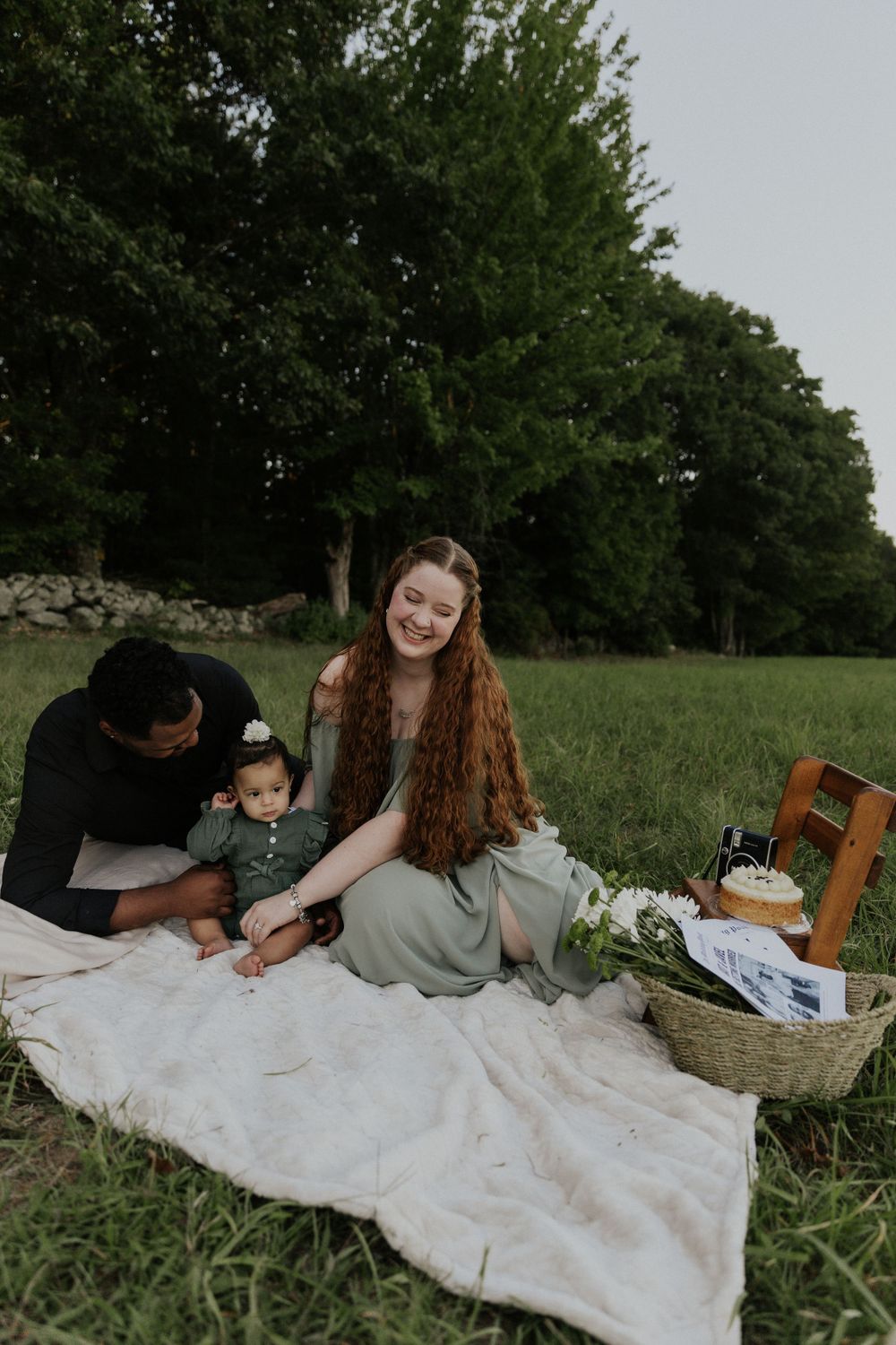 A family enjoys a relaxing picnic on a white blanket in a grassy field surrounded by trees.