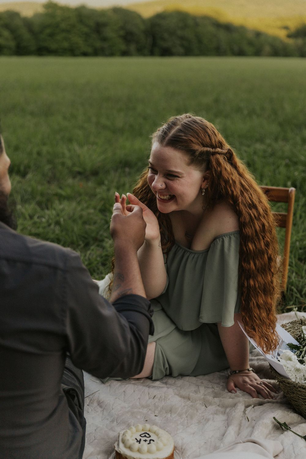 A romantic couple shares a candid moment while lying on a blanket in a grassy field at sunset.