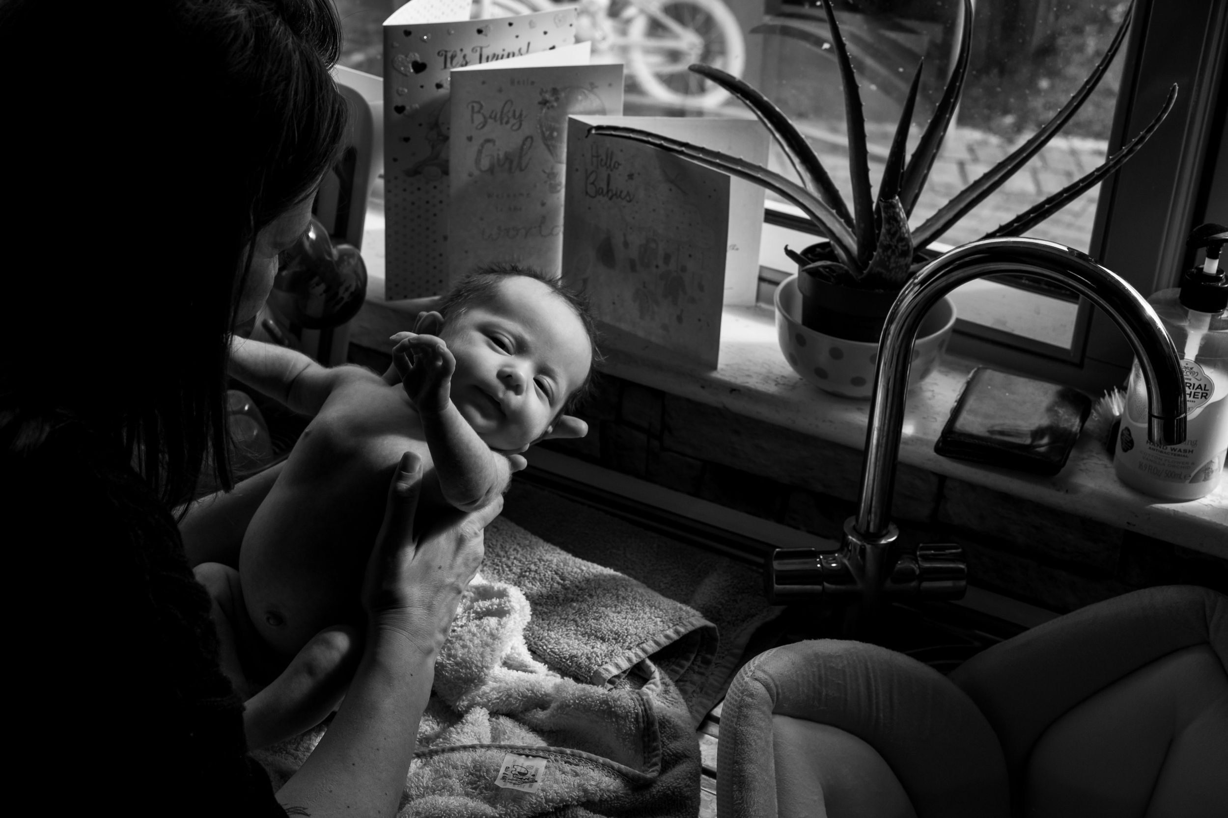 A small baby is being fed in a darkly lit room with a houseplant visible in the background.