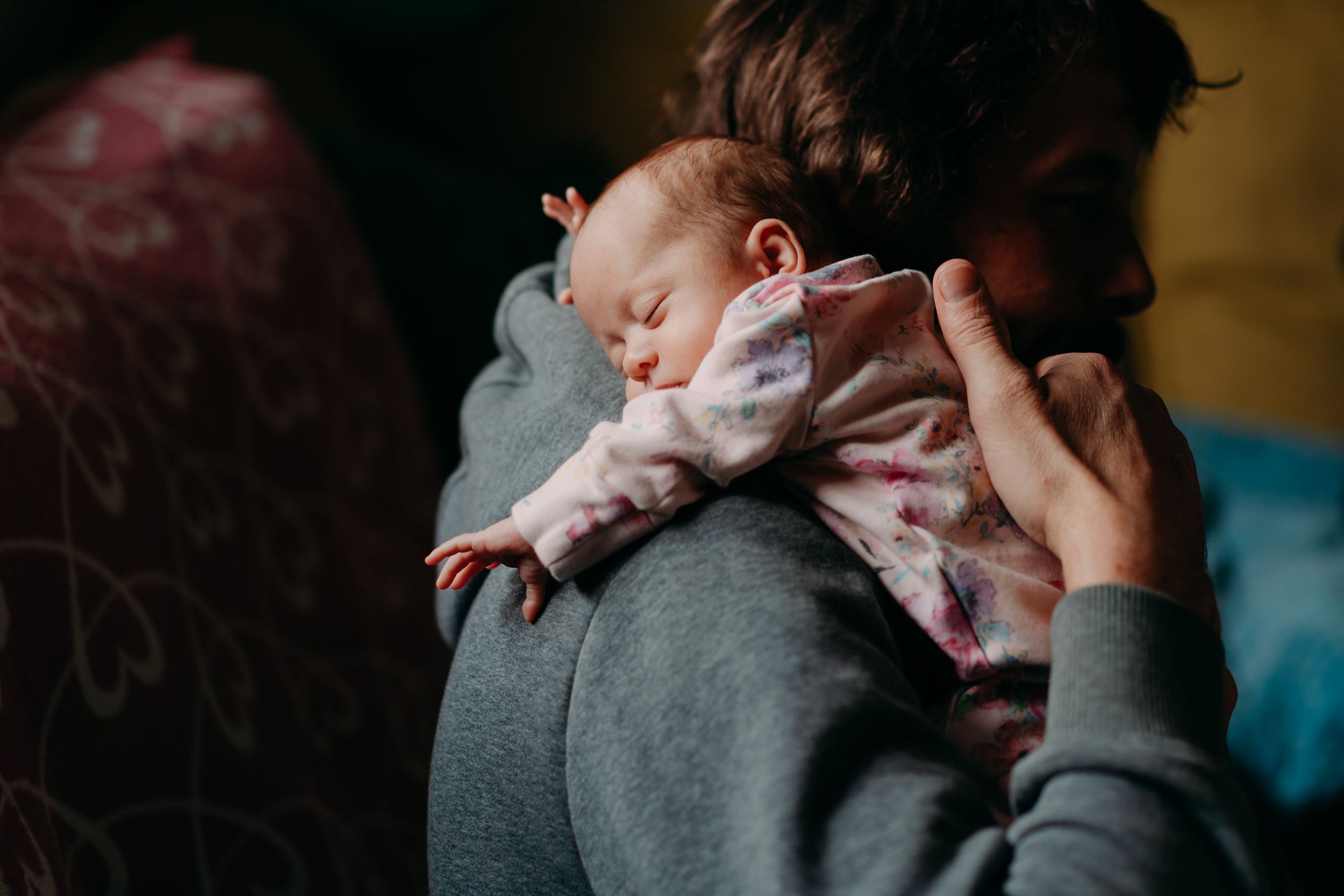 A sleeping baby in a pink outfit rests peacefully on someone wearing a gray sweater.