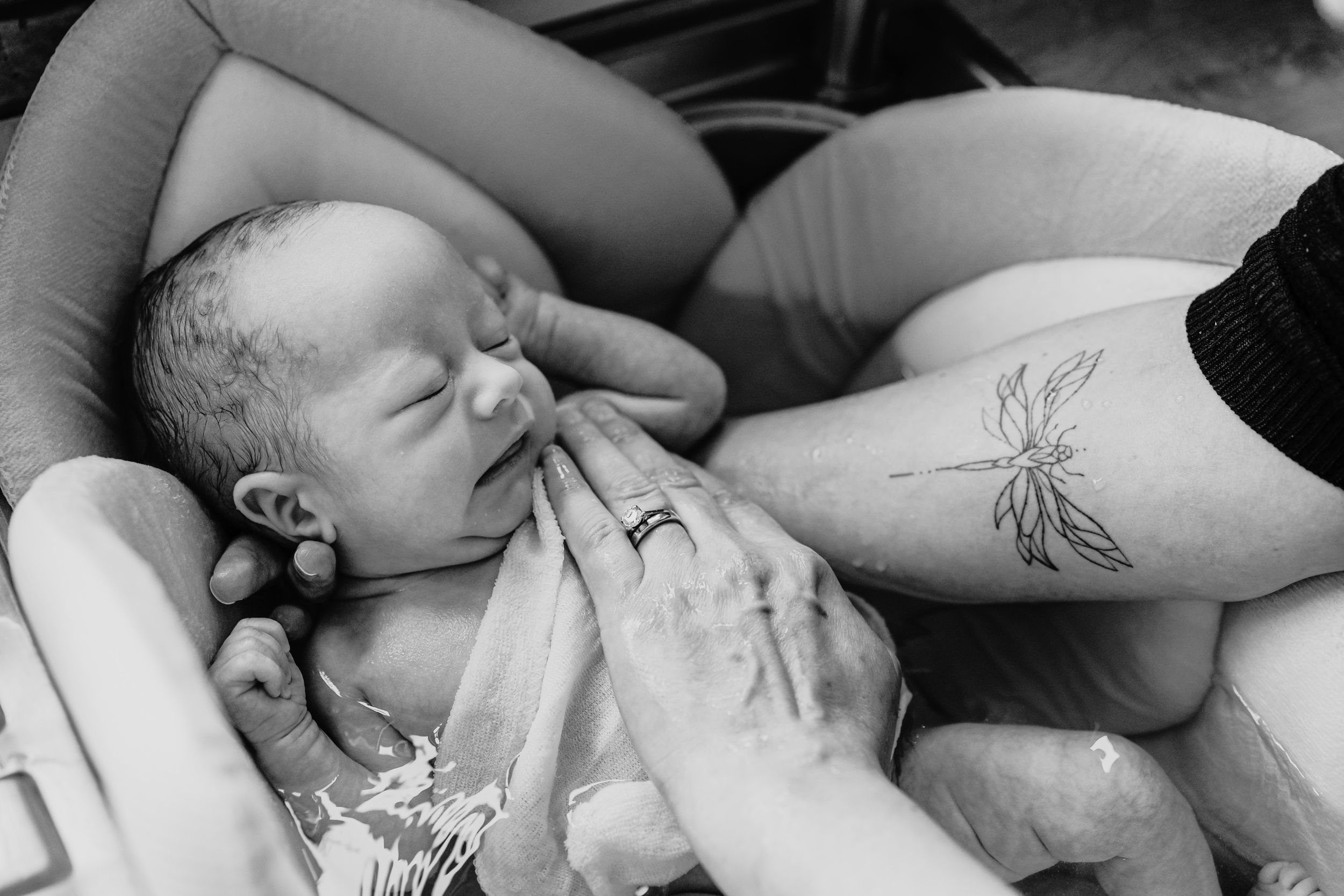 Black and white close up of newborn sleeping peacefully while resting against arm with dragonfly tattoo.