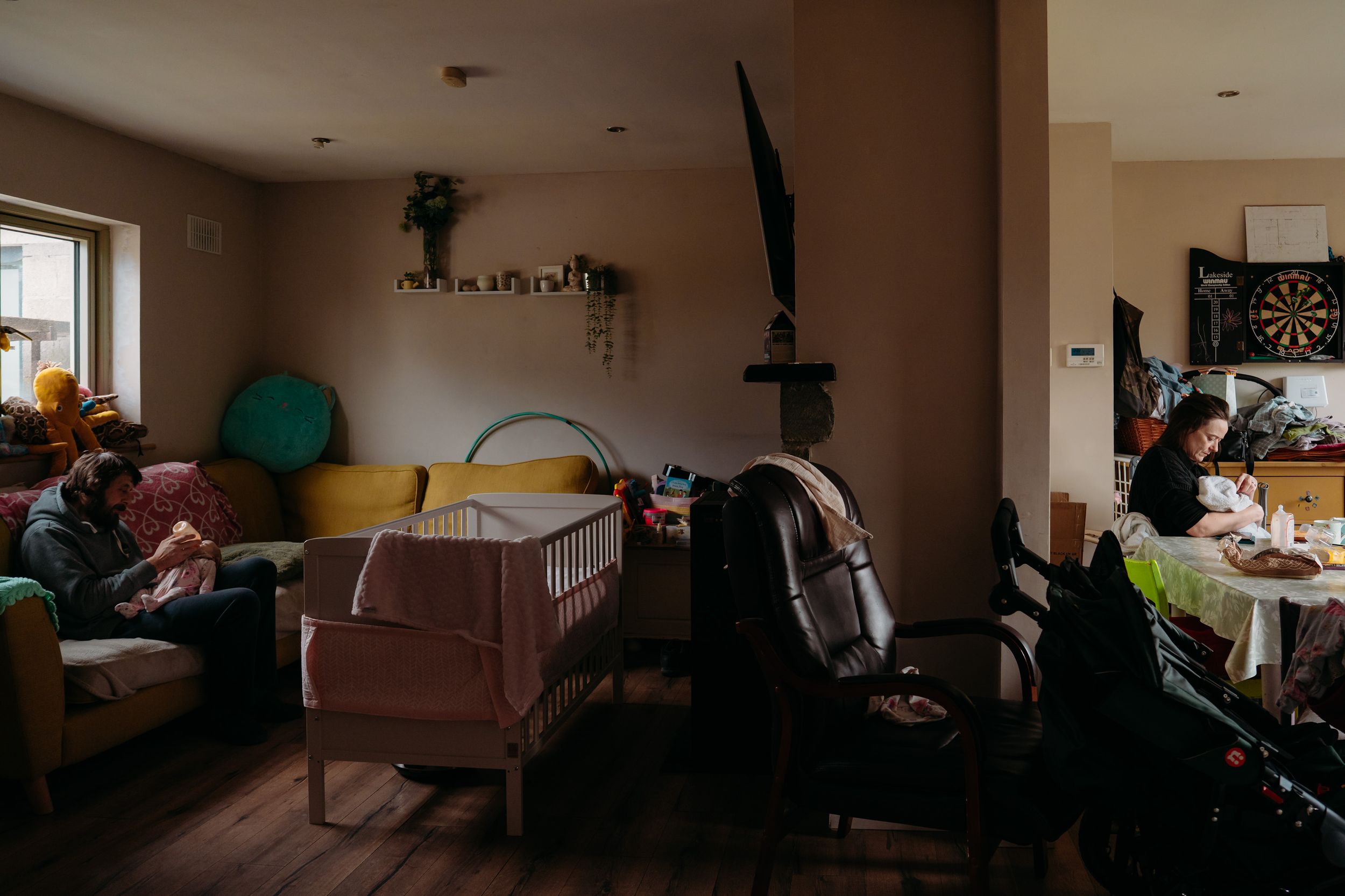 Mother feeds baby in the kitchen while Dad feeds her twin sister in the living room.
