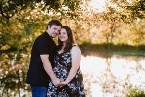 Maternity photo of a husband and wife smiling against a peaceful water backdrop.