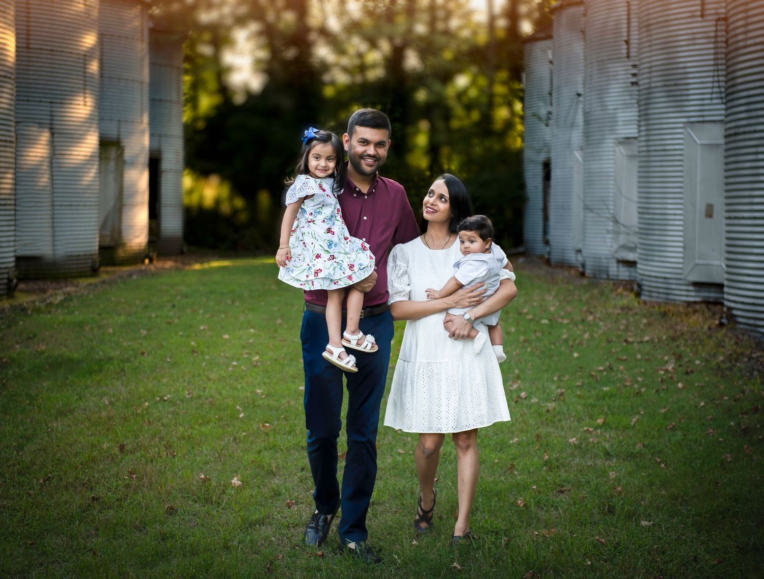 Young family poses together on grass near white industrial structures at sunset.