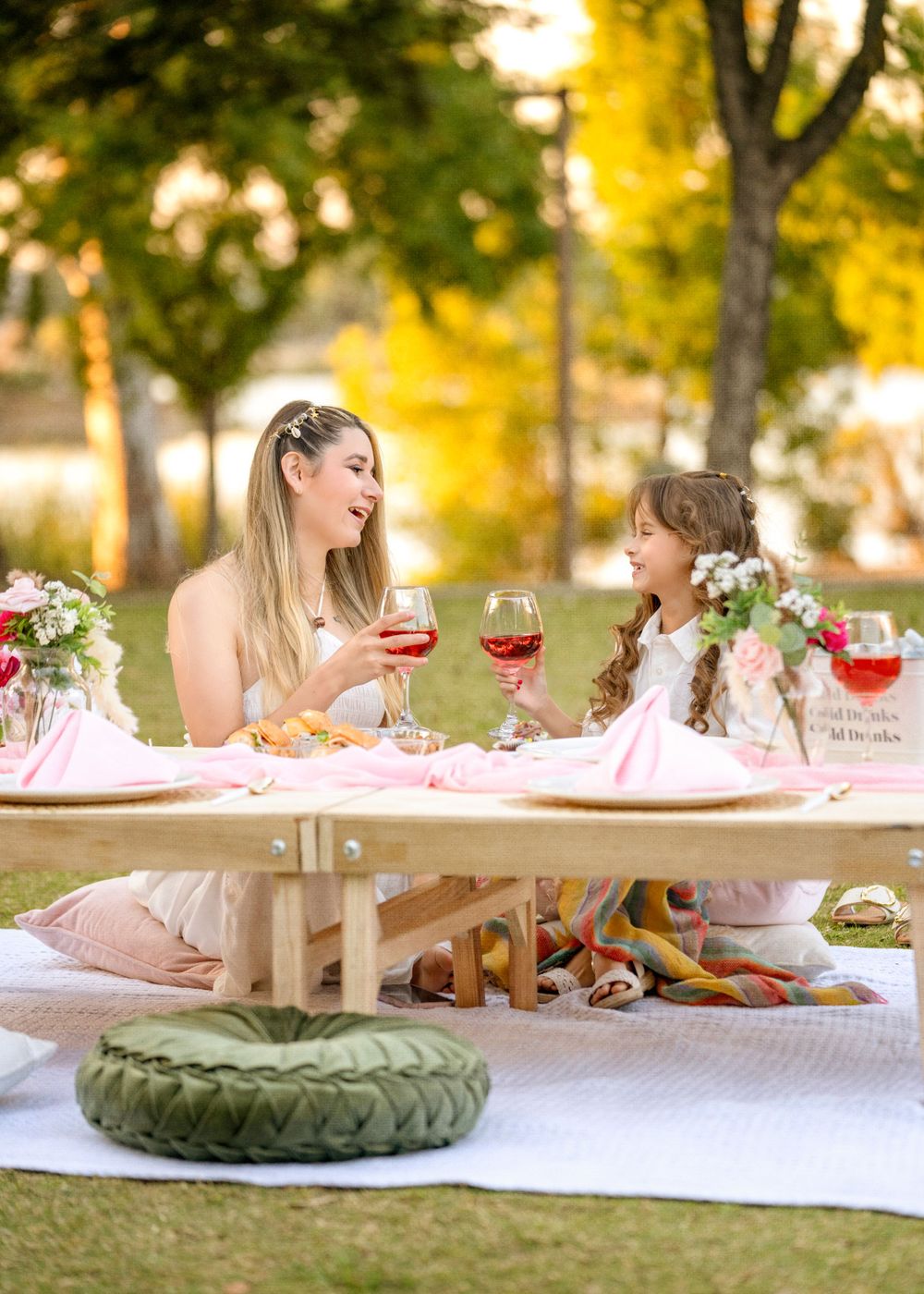Two people share a tea party picnic with wine glasses on a blanket outdoors at sunset.