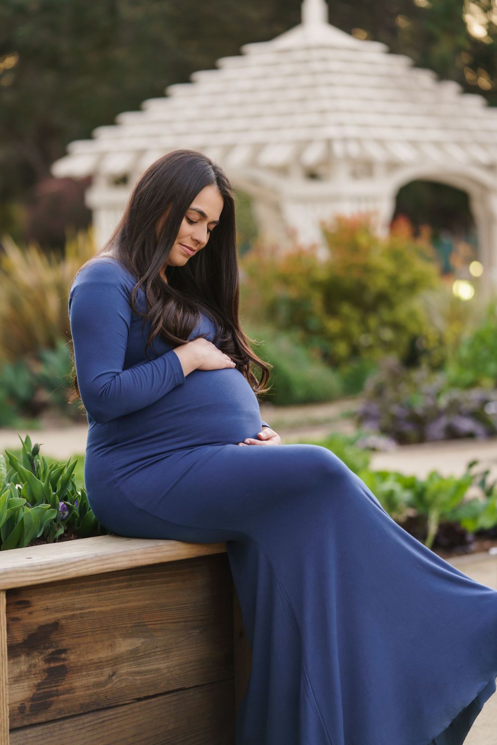 A maternity photo of an expectant mother in a flowing navy blue dress sitting in a garden setting with a white gazebo.