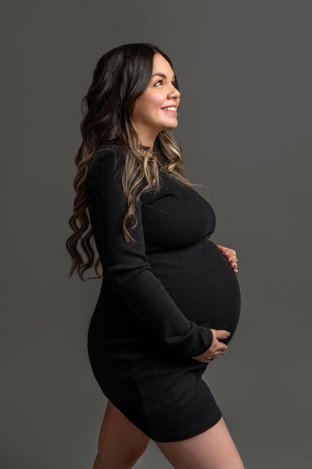 Side profile of an expectant mother wearing a black sweater dress against a gray backdrop.