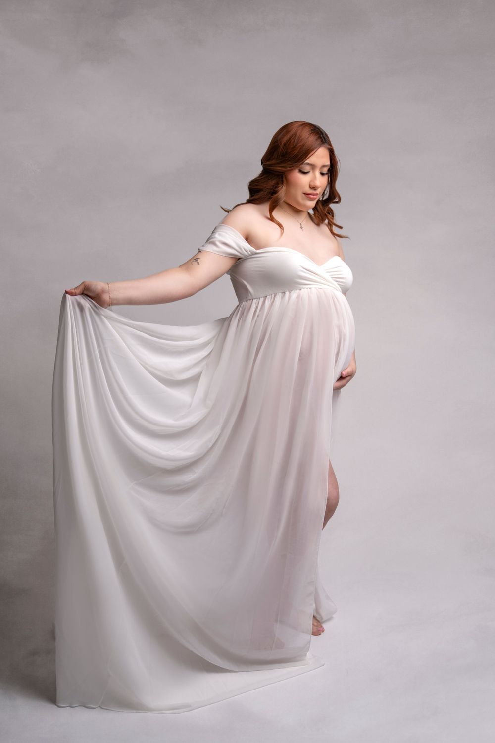 A maternity portrait featuring a flowing white gown against a gray studio backdrop.