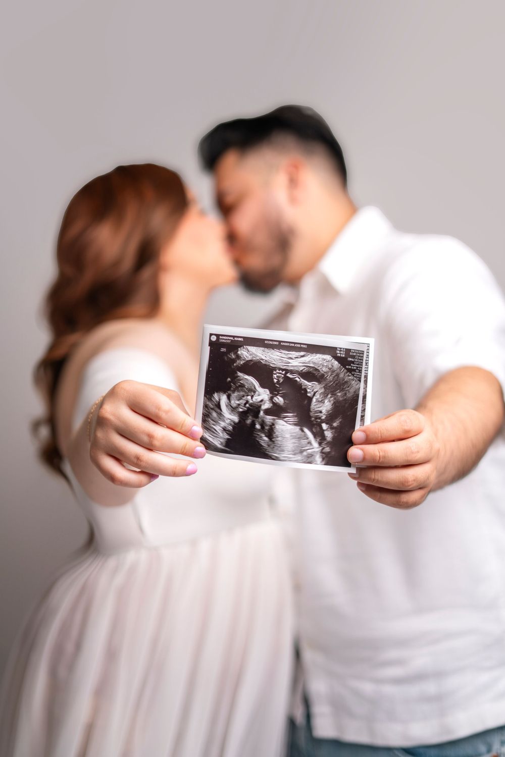 A couple shares an intimate moment while holding up their ultrasound photo against a gray background.
