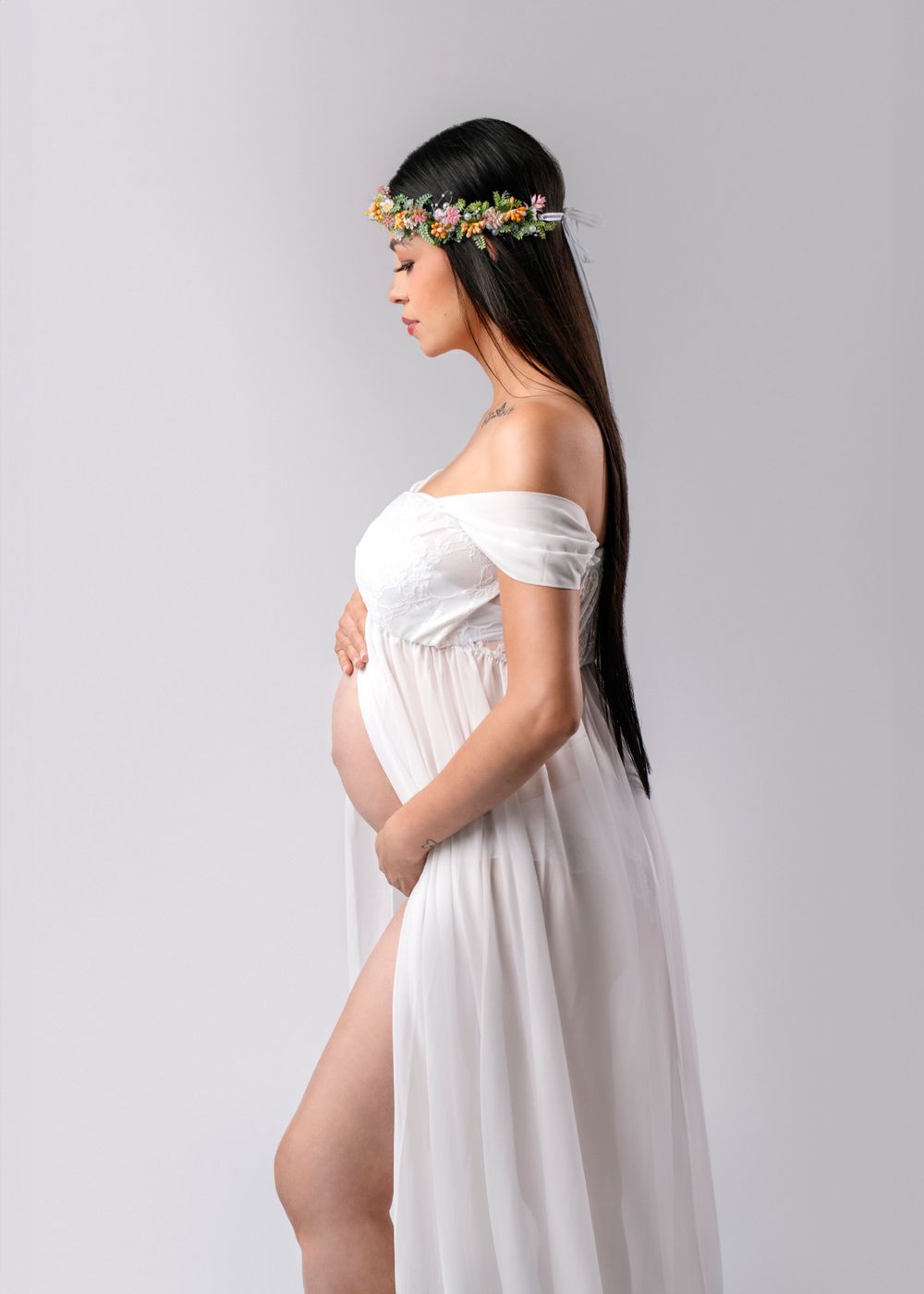 Side profile of expectant woman in white dress wearing floral crown against grey background.