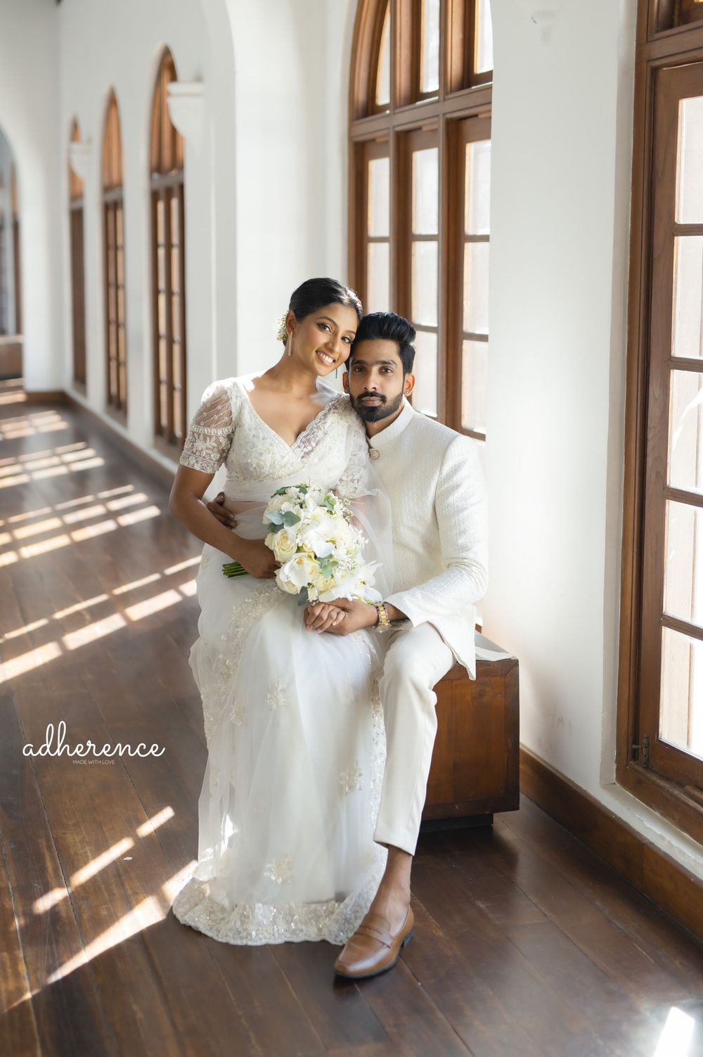 Elegant wedding couple in white attire pose in sunlit corridor with arched windows.