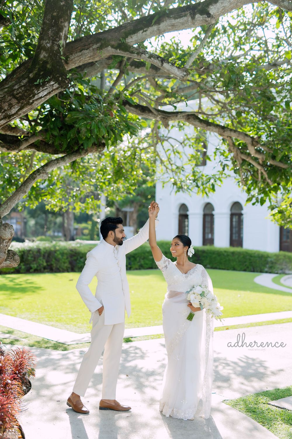 Joyful wedding couple in white outfits pose playfully under tree branches.