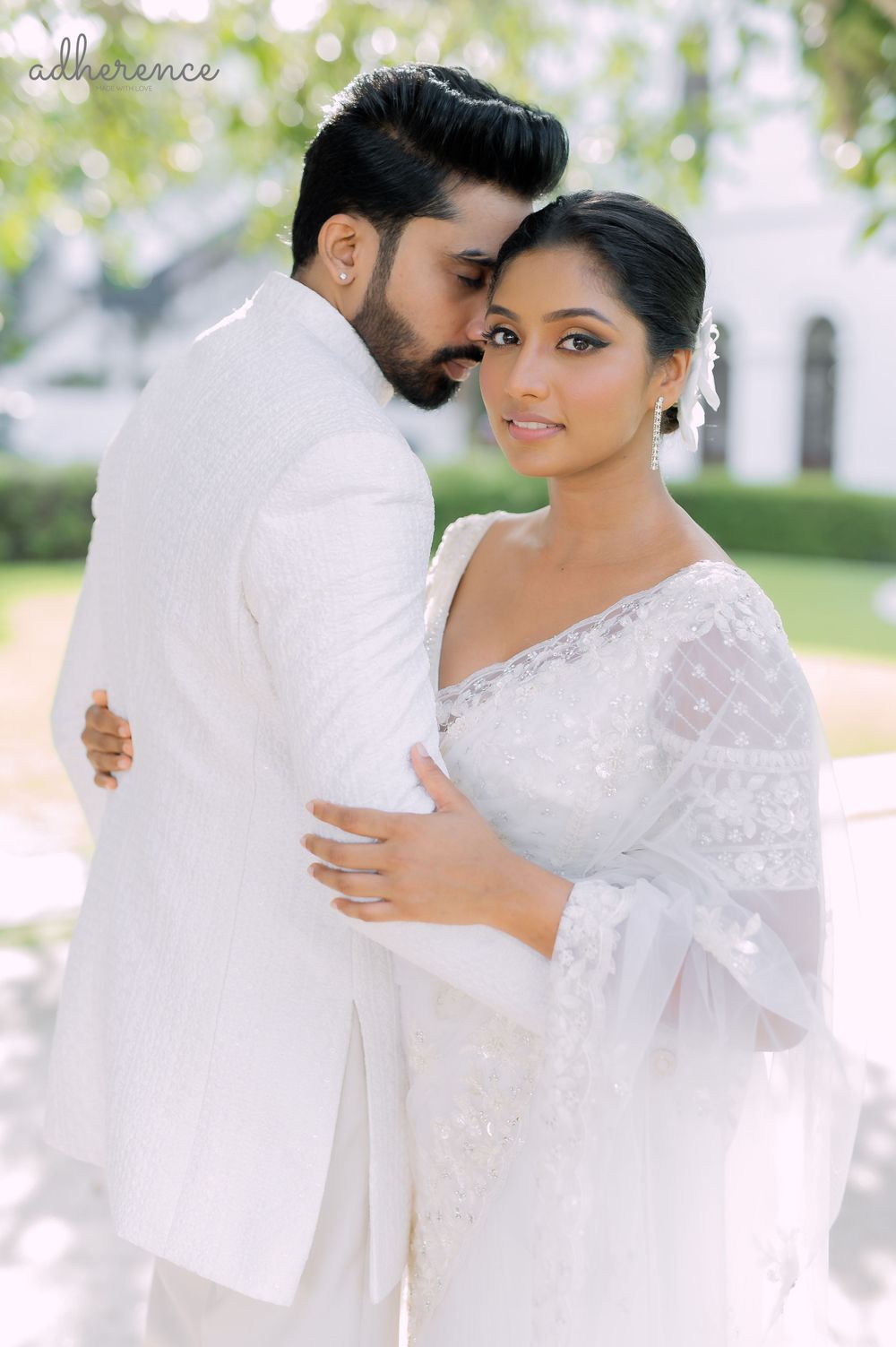 Intimate wedding portrait of couple in white traditional attire against garden backdrop.
