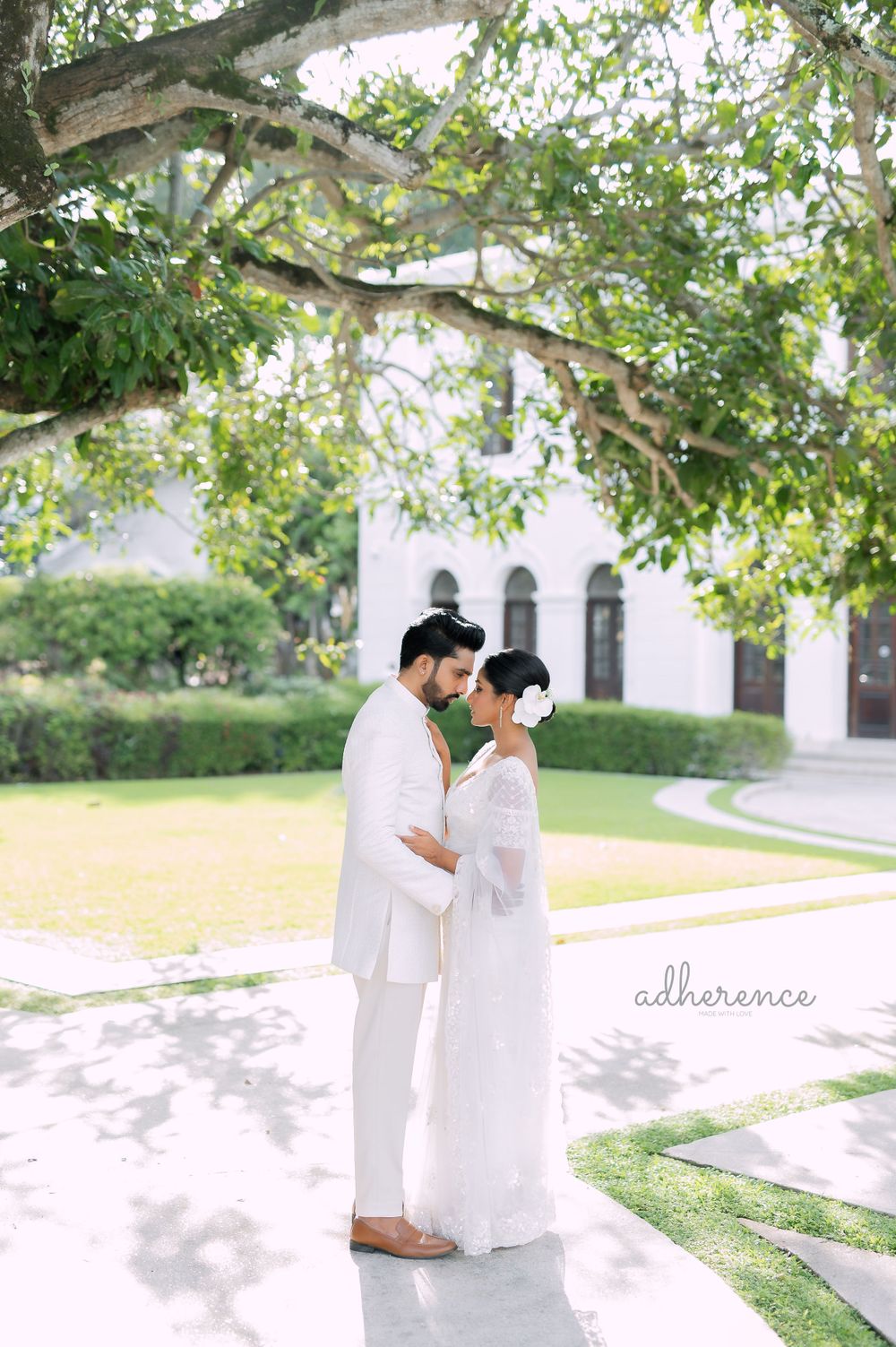 A couple in white traditional attire embraces outdoors near a historic building under tree branches.