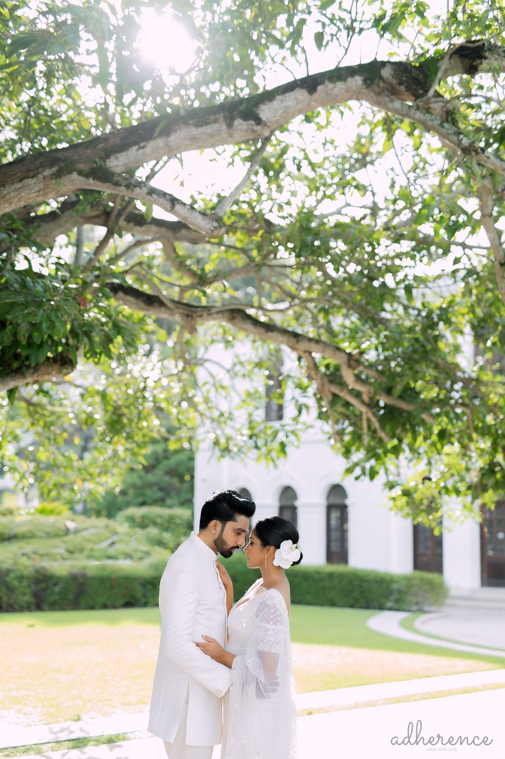 A romantic outdoor wedding portrait beneath sunlit tree branches and white building.