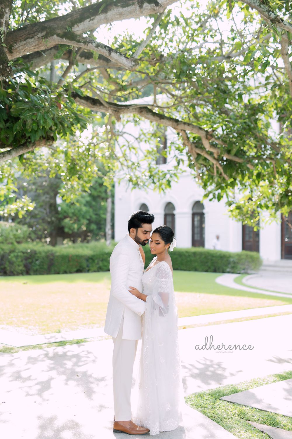 A couple shares an intimate moment outdoors under green trees near a white building.