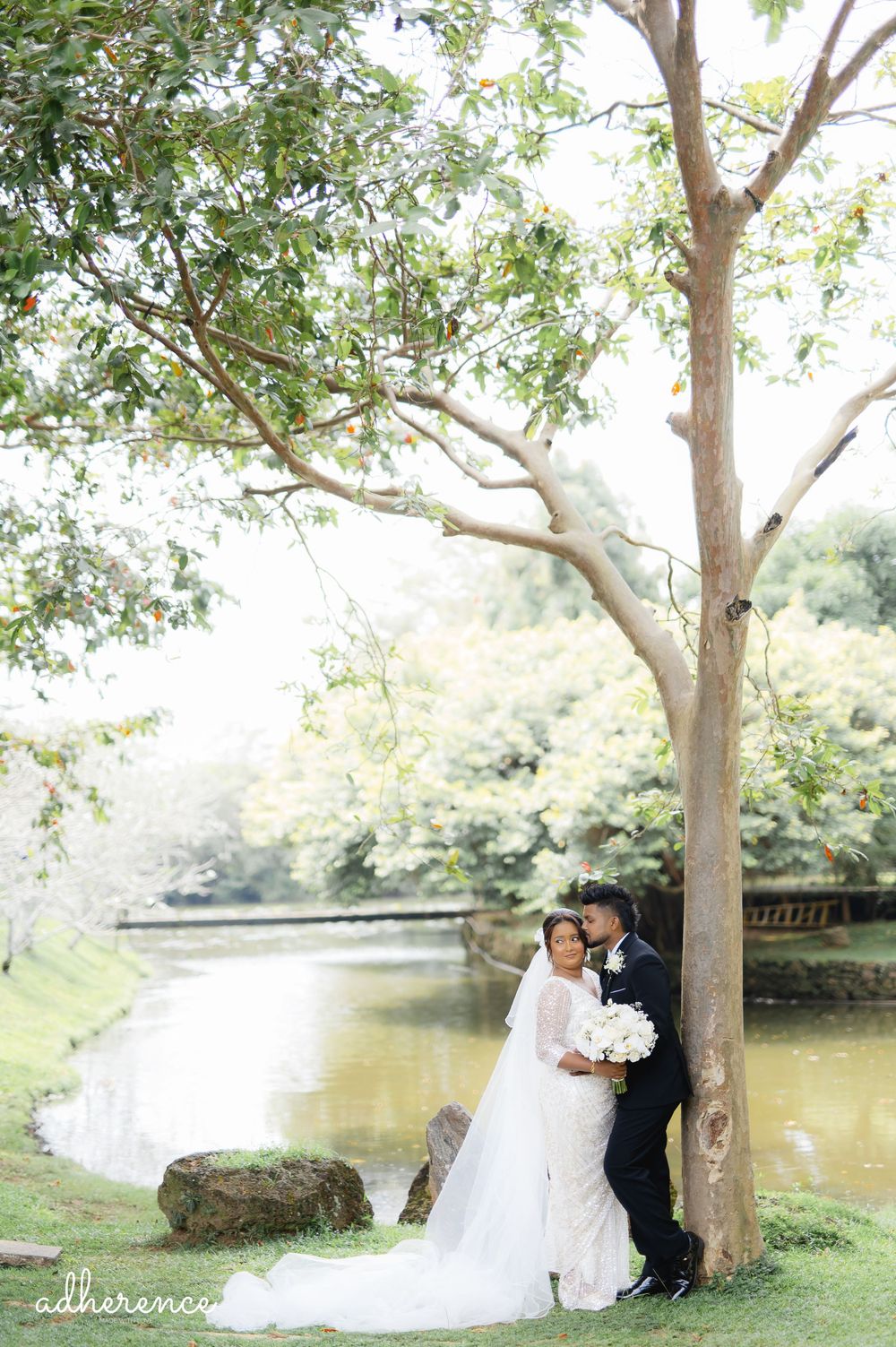 A romantic wedding photography moment captured by a lake with a large tree and lush greenery in the background.