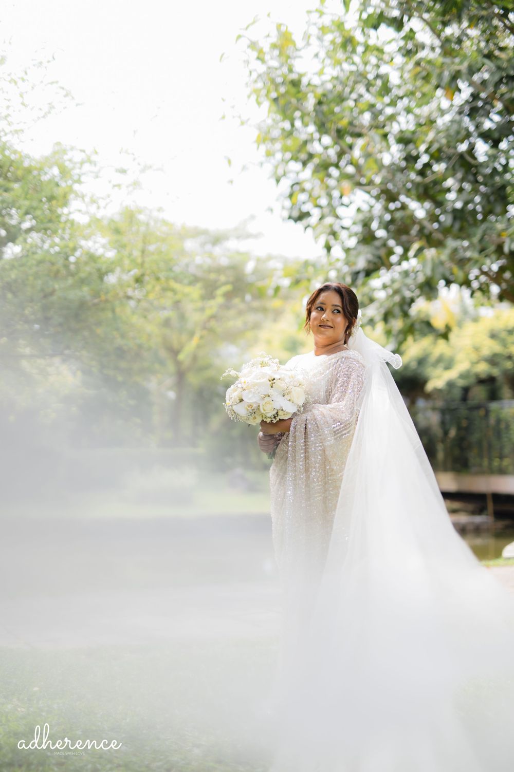Bride in a flowing white lace dress holds a white bouquet in a dreamy outdoor garden setting with misty lighting.