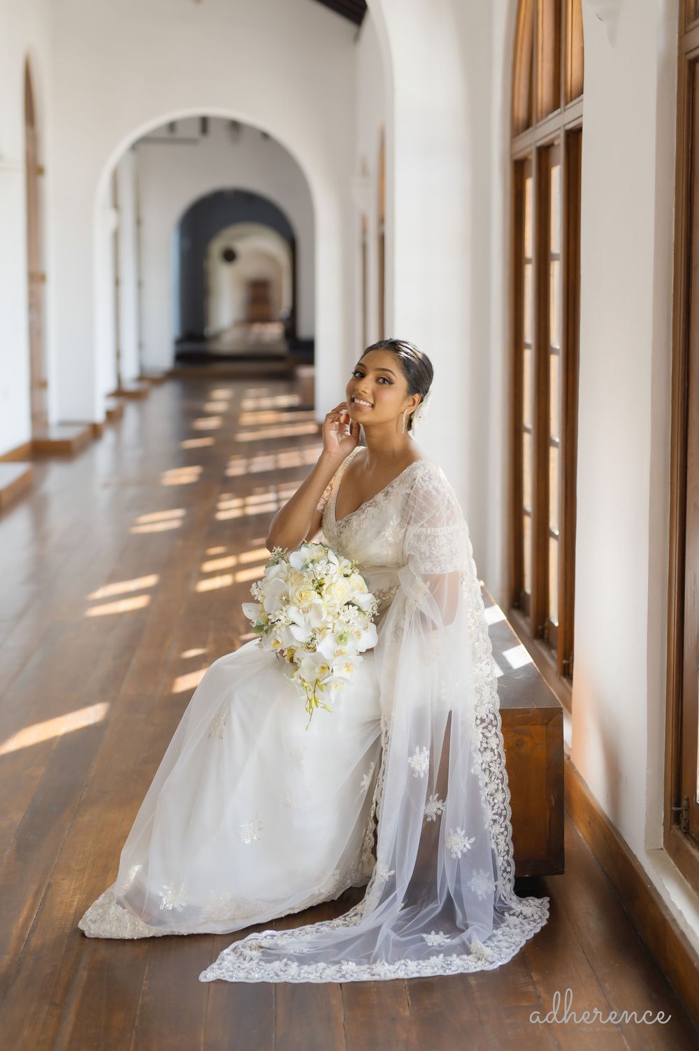 A bride in white lace gown holds bouquet while sitting in a sunlit corridor.