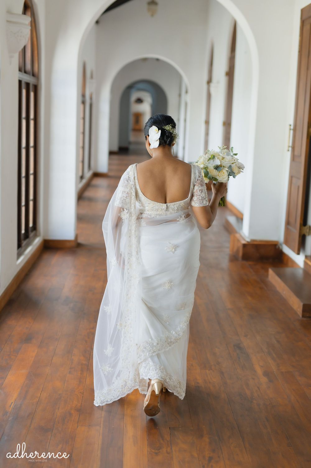 A bride walks down a hallway holding white flowers, showing the back detail of her lace wedding dress.
