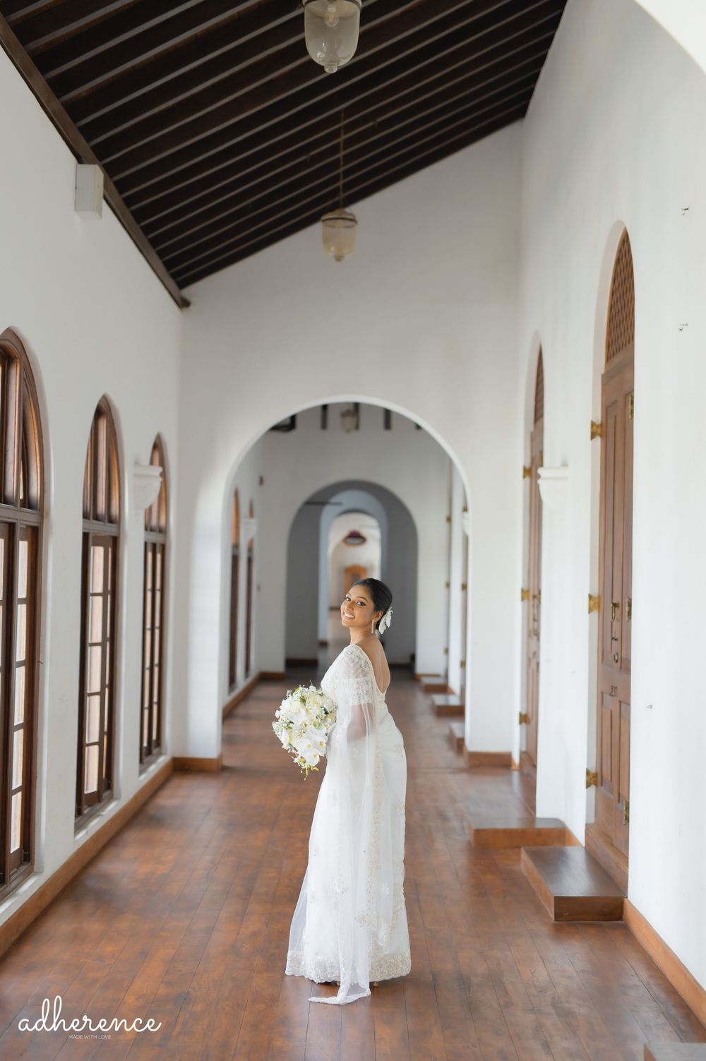 A bride poses with bouquet in a historic hallway with wooden ceiling and arched windows.
