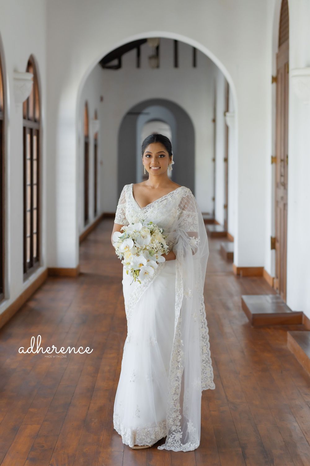 A bride in white lace gown poses with bouquet in an elegant arched hallway.