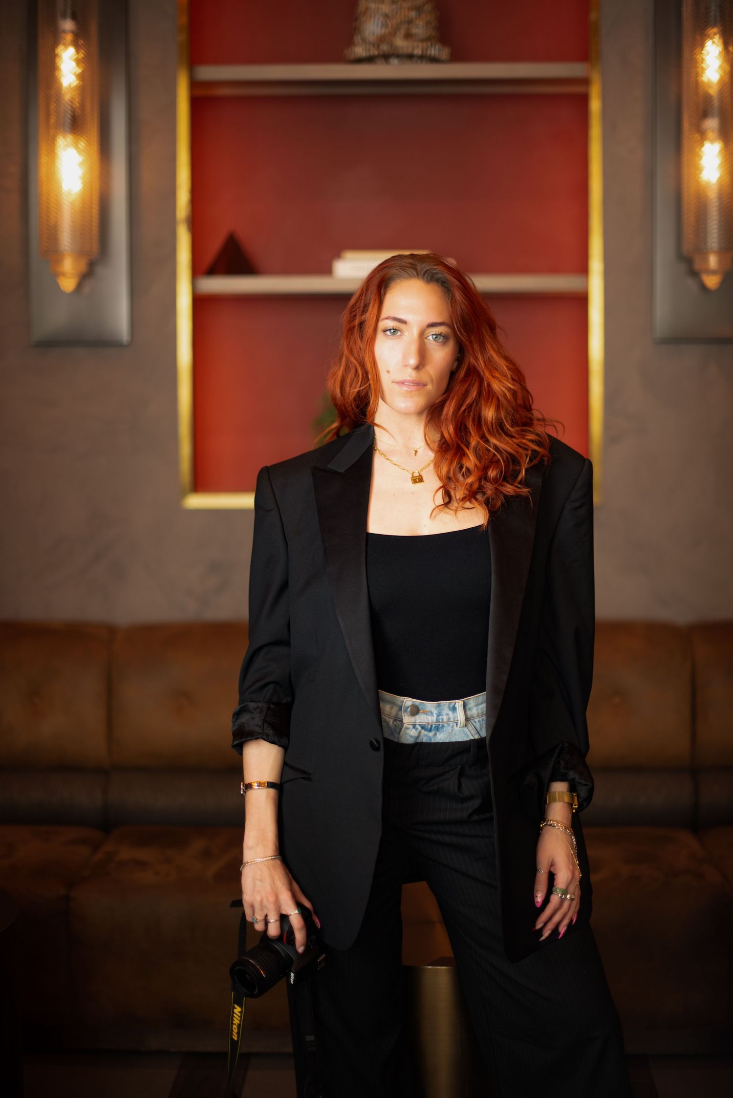Professional studio portrait of a person with red hair wearing a black blazer against a warm red wall with dramatic lighting.