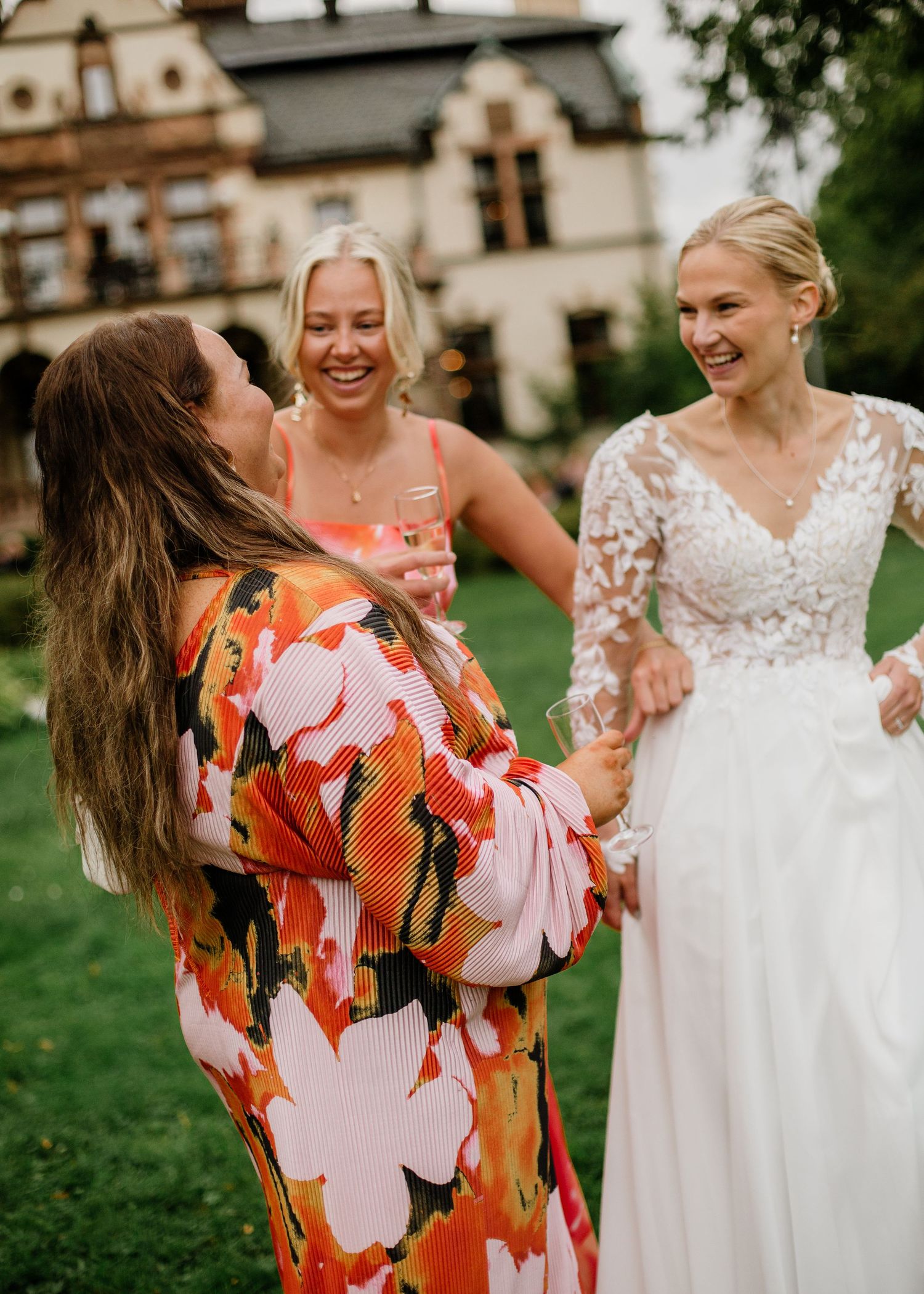 Bride in white lace gown laughing with guests in colorful dresses outside elegant manor house.