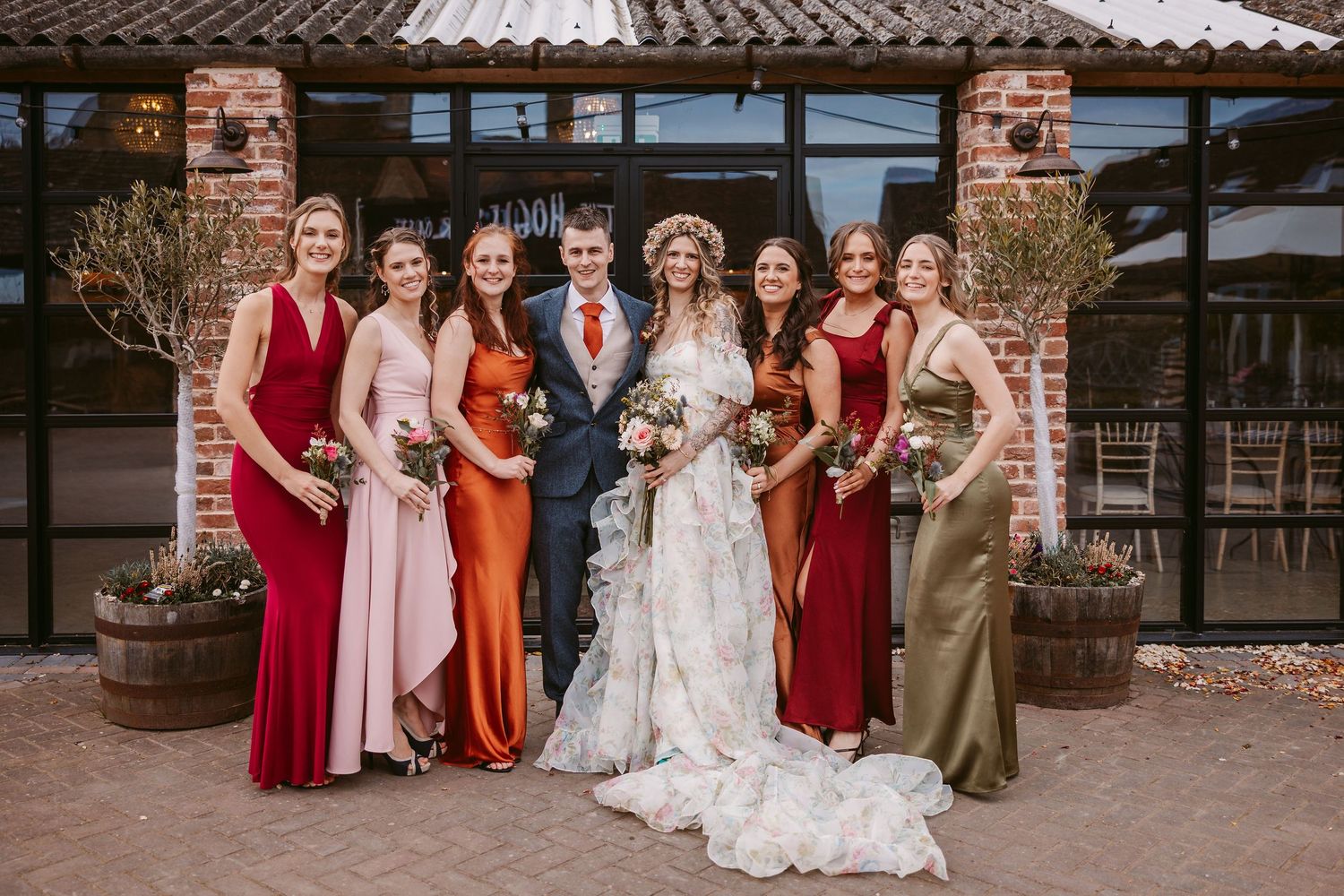 Wedding party in mix of red and neutral dresses poses together in front of brick wall at Stratton Court Barn wedding venue.