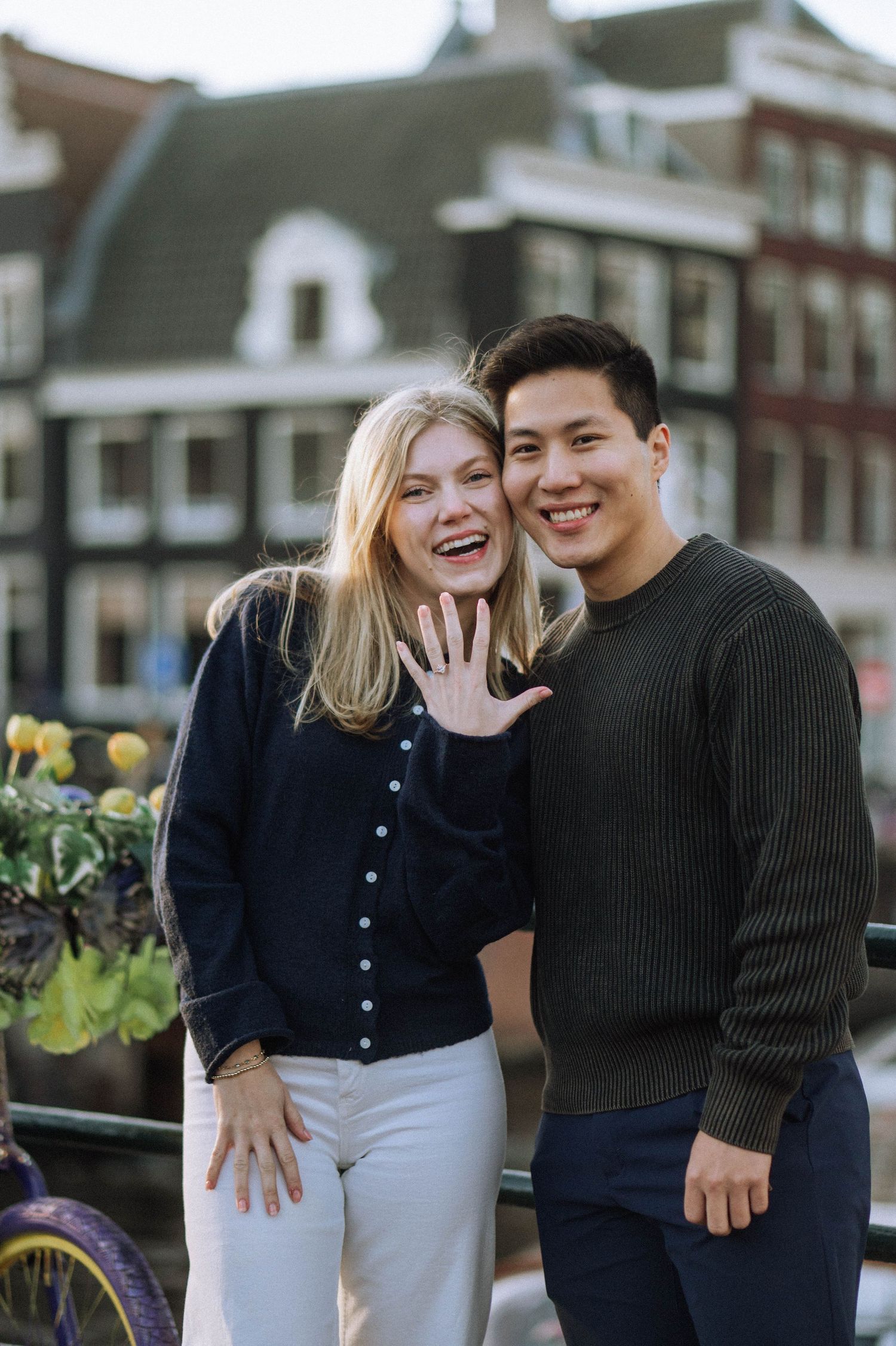 Happy couple laughing and showing engagement ring in urban Amsterdam setting with spring flowers in background.