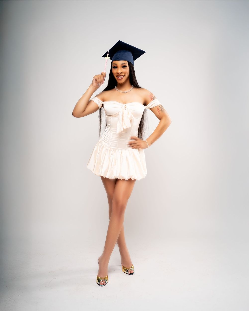 Graduate posing in white off-shoulder mini dress and graduation cap against white background.